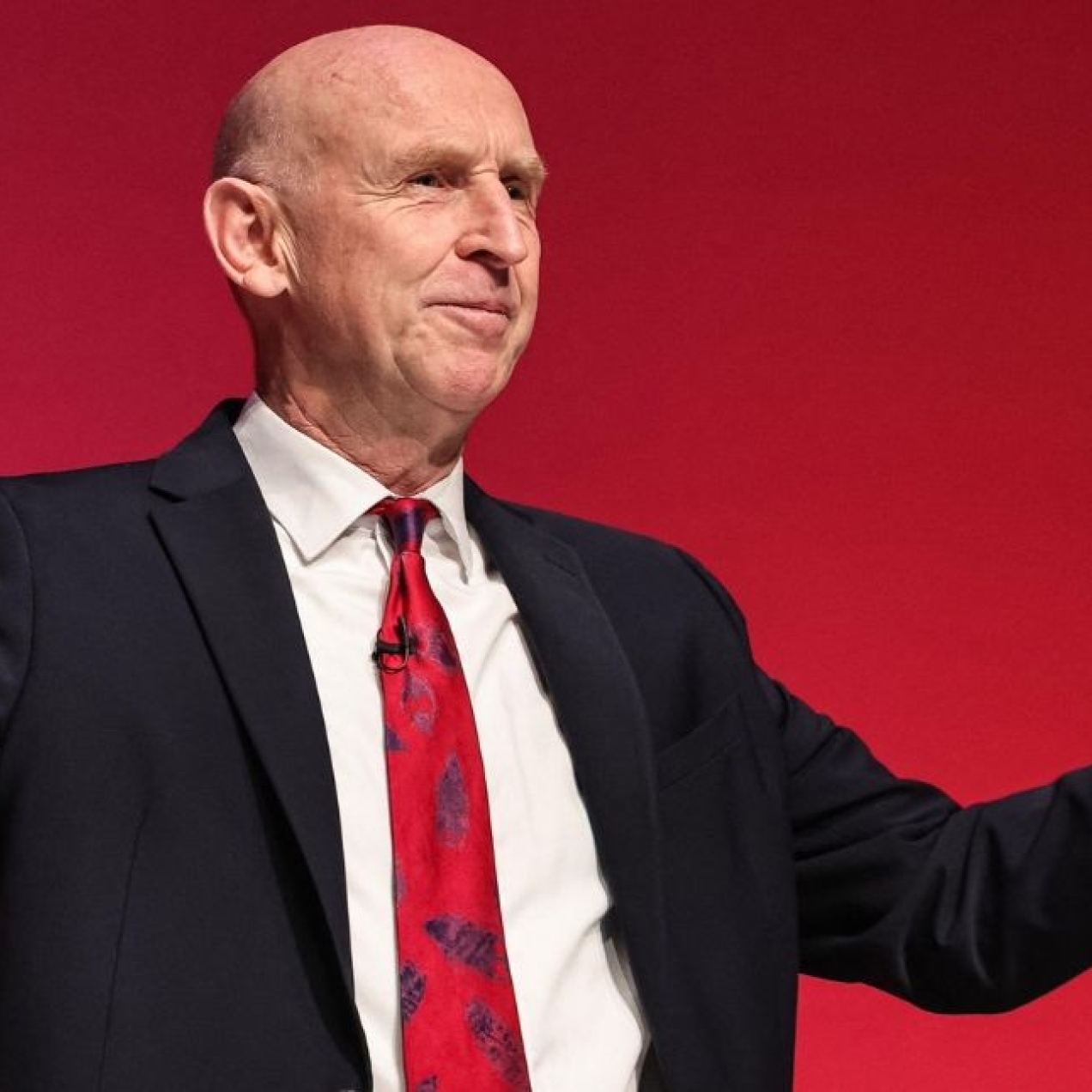 Chancellor John Healey - a white man wearing a dark suit - stood against a red background with his arms open wide.